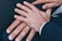Several hands are placed together, displaying wedding rings on their fingers. The hands appear to be resting on a dark fabric surface and are wearing formal attire.