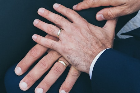 Several hands are placed together, displaying wedding rings on their fingers. The hands appear to be resting on a dark fabric surface and are wearing formal attire.