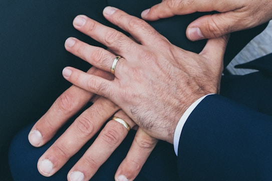 Several hands are placed together, displaying wedding rings on their fingers. The hands appear to be resting on a dark fabric surface and are wearing formal attire.