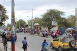 A busy urban street scene with people walking along the sidewalk and wearing pink shirts. A yellow vehicle and motorcycles are present on the road. A university building is visible in the background with a large tree nearby and a sign indicating its name. Several market stalls and umbrellas add to the vibrant atmosphere.