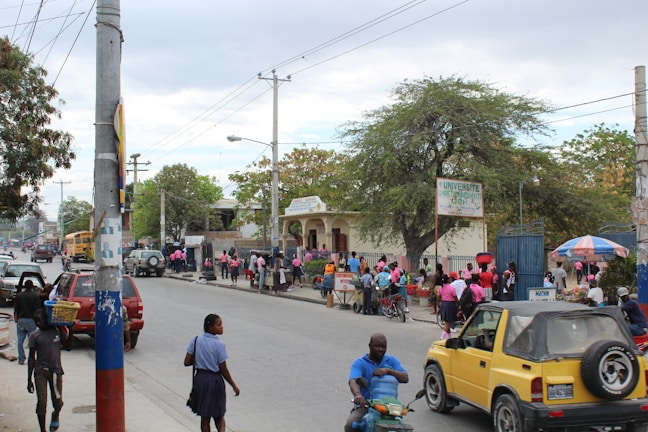 A busy urban street scene with people walking along the sidewalk and wearing pink shirts. A yellow vehicle and motorcycles are present on the road. A university building is visible in the background with a large tree nearby and a sign indicating its name. Several market stalls and umbrellas add to the vibrant atmosphere.
