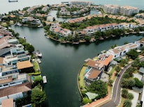 Aerial view of a residential area with modern houses surrounded by a winding body of water. The community features greenery, docks, and boats along the canal. This upscale neighborhood is situated by a larger water body, likely near a coastal area, with additional buildings in the background.