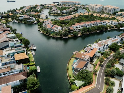 Aerial view of a residential area with modern houses surrounded by a winding body of water. The community features greenery, docks, and boats along the canal. This upscale neighborhood is situated by a larger water body, likely near a coastal area, with additional buildings in the background.