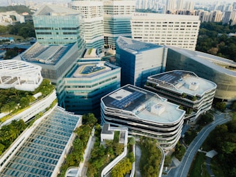 Aerial view of a modern urban complex featuring multiple futuristic-style buildings with glass facades and rooftop gardens. The architecture is characterized by sleek, geometric designs. Surrounding the area are patches of green spaces and a network of roads. In the background, a city skyline is visible.