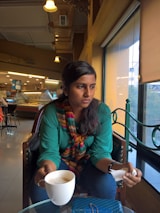 A person sits in a cafe holding a cup of coffee, looking pensively out of a large window. The cafe interior is warmly lit with decorative light fixtures, and several tables and chairs are visible in the background. The person is wearing a green shirt with a colorful scarf, and a blue purse rests on the table in front.