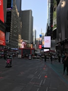 A bustling urban scene with tall skyscrapers lining the streets. Numerous electronic billboards and advertisements are prominently visible. People are walking or standing around in the open space, with some seated at tables. The environment reflects a lively city atmosphere.