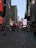A bustling urban scene with tall skyscrapers lining the streets. Numerous electronic billboards and advertisements are prominently visible. People are walking or standing around in the open space, with some seated at tables. The environment reflects a lively city atmosphere.