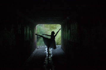 A silhouette of a dancer striking a graceful pose inside a dimly lit tunnel. The light at the end of the tunnel highlights the dancer's form, creating a dramatic and artistic effect. The background outside the tunnel appears lush and green, suggesting a natural environment.