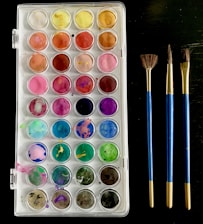 A palette of watercolor paints consisting of multiple compartments with a range of vibrant colors is arranged neatly. Beside it, there are three paintbrushes with blue handles and gold ferrules placed parallel to each other. The palette includes colors such as yellow, orange, red, pink, purple, blue, green, and brown. The background is dark, contrasting with the bright colors of the paints.
