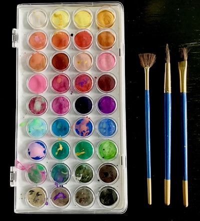 A palette of watercolor paints consisting of multiple compartments with a range of vibrant colors is arranged neatly. Beside it, there are three paintbrushes with blue handles and gold ferrules placed parallel to each other. The palette includes colors such as yellow, orange, red, pink, purple, blue, green, and brown. The background is dark, contrasting with the bright colors of the paints.