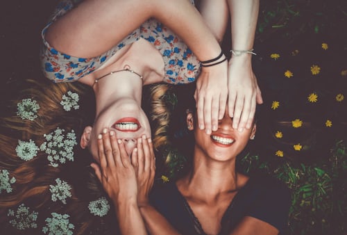 Twee jonge vrouwen die naast elkaar op het gras liggen en elkaars ogen bedekken voor een Instagram-foto. 