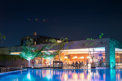 A modern outdoor setting featuring a swimming pool reflecting the night lights from a nearby building, which has a glass facade revealing people dining inside. Trees are illuminated with bright lights, creating a vibrant atmosphere.