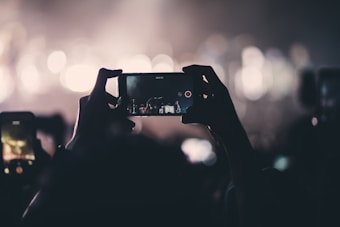 A person holds up a smartphone, capturing a video or image of a live event in a dimly lit environment. The background is filled with bright, blurred lights that create a bokeh effect, giving a sense of a concert or performance setting. The foreground features silhouettes of other people holding up their phones as well.