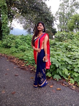 A woman stands on a rural road surrounded by lush greenery, wearing a traditional red and blue sari with gold embellishments. She holds a phone in her hand and smiles at the camera.
