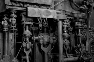 A complex assembly of mechanical components featuring large metal rods, pistons, and gears, indicating a part of industrial machinery. The structure is weathered and worn, with visible signs of rust and age. Thick cables and pipes run alongside the metal parts, contributing to a sophisticated and intricate design.