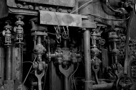 A complex assembly of mechanical components featuring large metal rods, pistons, and gears, indicating a part of industrial machinery. The structure is weathered and worn, with visible signs of rust and age. Thick cables and pipes run alongside the metal parts, contributing to a sophisticated and intricate design.
