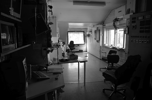 A dimly lit, abandoned control room with multiple monitors and control panels visible. The room is cluttered with old computers, keyboards, and papers scattered across the desks. Chairs are positioned haphazardly, and cables hang loosely from the edges. The windows allow some natural light into the room, illuminating the dust particles in the air.