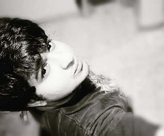 A black-and-white portrait of a person looking upwards, capturing detailed facial features and textured hair. The photo is taken at an overhead angle, emphasizing the expression and mood of the subject.