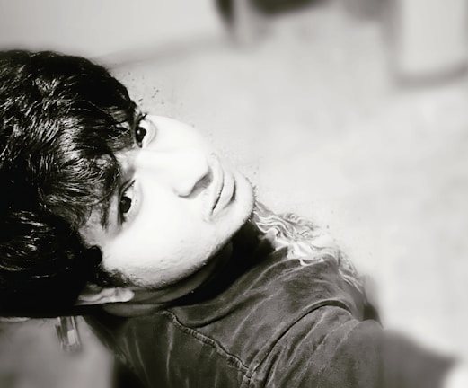 A black-and-white portrait of a person looking upwards, capturing detailed facial features and textured hair. The photo is taken at an overhead angle, emphasizing the expression and mood of the subject.