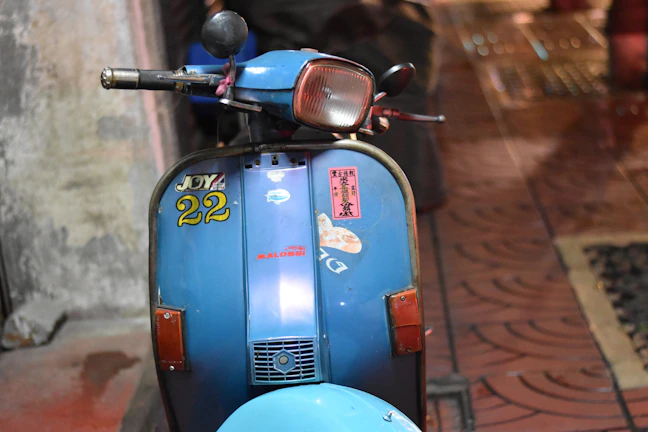 A vintage blue scooter with various stickers and decals is parked against a textured wall. The scooter features a central headlight and a registration number, 'JOY 22', displayed prominently. The setting appears to be an indoor space with tiled flooring and warm lighting.
