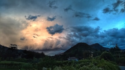 A dramatic sky filled with dark and brooding clouds transitions into a vibrant sunset with shades of orange and blue. The landscape below features rolling hills and lush greenery, with a few houses scattered in the foreground. A single street lamp is visible against the backdrop of the hilly terrain.