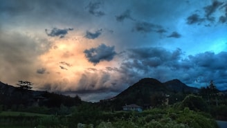 A dramatic sky filled with dark and brooding clouds transitions into a vibrant sunset with shades of orange and blue. The landscape below features rolling hills and lush greenery, with a few houses scattered in the foreground. A single street lamp is visible against the backdrop of the hilly terrain.