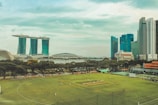 A lush green cricket field is in the foreground with players dressed in white visibly engaged in a match. Surrounding the field are tall trees and some red-roofed structures. Behind the field, a river can be seen with an iconic, uniquely designed building with three towers joined at the top by a horizontal structure, and several modern skyscrapers. The sky above is partly cloudy with hints of blue.