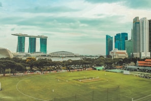 A lush green cricket field is in the foreground with players dressed in white visibly engaged in a match. Surrounding the field are tall trees and some red-roofed structures. Behind the field, a river can be seen with an iconic, uniquely designed building with three towers joined at the top by a horizontal structure, and several modern skyscrapers. The sky above is partly cloudy with hints of blue.
