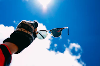 A close-up of sunglasses reflecting a bright sun and blue sky, held in hand.