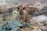 A small brown and black puppy sits amidst a pile of trash and debris, which includes plastic bags, a tattered green cloth, and other discarded items. The background appears to be a cluttered and possibly polluted environment.