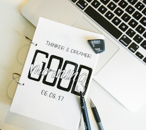 A close-up of a notebook with the words 'Thinker & Dreamer' and 'Good Morning' printed on it, dated 06.08.17. The notebook is placed next to an open laptop, and a black pen and mechanical pencil are lying beside it. An eraser is also visible.