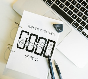 A close-up of a notebook with the words 'Thinker & Dreamer' and 'Good Morning' printed on it, dated 06.08.17. The notebook is placed next to an open laptop, and a black pen and mechanical pencil are lying beside it. An eraser is also visible.