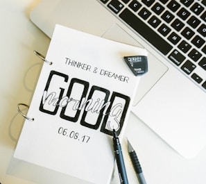 A close-up of a notebook with the words 'Thinker & Dreamer' and 'Good Morning' printed on it, dated 06.08.17. The notebook is placed next to an open laptop, and a black pen and mechanical pencil are lying beside it. An eraser is also visible.