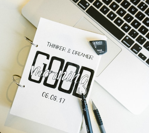 A close-up of a notebook with the words 'Thinker & Dreamer' and 'Good Morning' printed on it, dated 06.08.17. The notebook is placed next to an open laptop, and a black pen and mechanical pencil are lying beside it. An eraser is also visible.