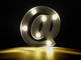 A glowing at symbol with embedded lights is placed on a surface, emitting a warm yellow-white glow against a dark background.