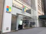 The storefront of a Microsoft retail store with large glass windows and the company's logo prominently displayed. A poster advertising the Surface Laptop is visible near the entrance. The architecture features a modern look with a clean facade and sleek design. A couple of people can be seen inside the store on the second floor.