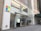 The storefront of a Microsoft retail store with large glass windows and the company's logo prominently displayed. A poster advertising the Surface Laptop is visible near the entrance. The architecture features a modern look with a clean facade and sleek design. A couple of people can be seen inside the store on the second floor.