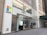 The storefront of a Microsoft retail store with large glass windows and the company's logo prominently displayed. A poster advertising the Surface Laptop is visible near the entrance. The architecture features a modern look with a clean facade and sleek design. A couple of people can be seen inside the store on the second floor.