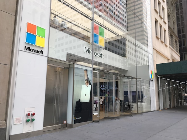 The storefront of a Microsoft retail store with large glass windows and the company's logo prominently displayed. A poster advertising the Surface Laptop is visible near the entrance. The architecture features a modern look with a clean facade and sleek design. A couple of people can be seen inside the store on the second floor.