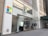 The storefront of a Microsoft retail store with large glass windows and the company's logo prominently displayed. A poster advertising the Surface Laptop is visible near the entrance. The architecture features a modern look with a clean facade and sleek design. A couple of people can be seen inside the store on the second floor.