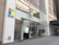 The storefront of a Microsoft retail store with large glass windows and the company's logo prominently displayed. A poster advertising the Surface Laptop is visible near the entrance. The architecture features a modern look with a clean facade and sleek design. A couple of people can be seen inside the store on the second floor.