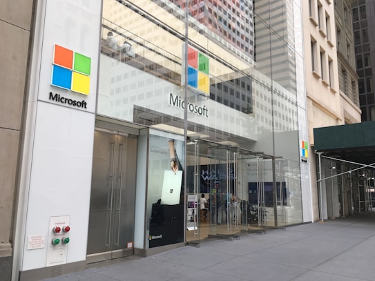 The storefront of a Microsoft retail store with large glass windows and the company's logo prominently displayed. A poster advertising the Surface Laptop is visible near the entrance. The architecture features a modern look with a clean facade and sleek design. A couple of people can be seen inside the store on the second floor.