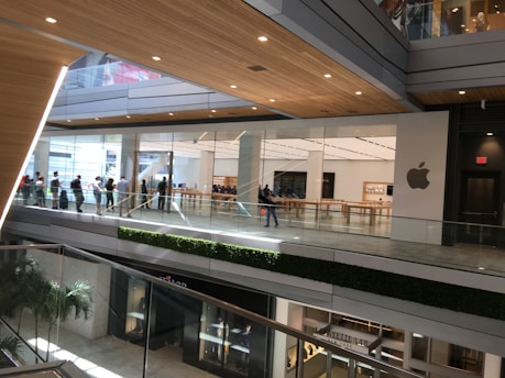 A modern, multi-level shopping mall with an Apple store featuring large glass windows and the Apple logo prominently displayed. People are seen walking and lining up along the upper floor. The space is well-lit with natural light, and there are elements of greenery on the balustrades.