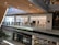 A modern, multi-level shopping mall with an Apple store featuring large glass windows and the Apple logo prominently displayed. People are seen walking and lining up along the upper floor. The space is well-lit with natural light, and there are elements of greenery on the balustrades.