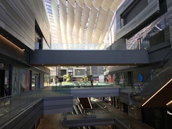 A modern, multi-level shopping mall interior with sleek architectural lines and ample natural light filtering through a unique, curvilinear ceiling design. The area includes pathways and glass railings with various shops visible along the sides. Decor includes modern advertisements and scattered potted plants.