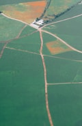 An aerial view of expansive green fields intersected by dirt roads leading to a white building surrounded by trees. The fields are divided into sections, some with visible patches of orange, possibly indicating seasonal changes or different crops.
