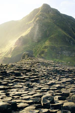 A landscape featuring a formation of interlocking basalt columns against the backdrop of a lush green mountain. The sunlight softly illuminates the mountainous terrain, creating a serene and majestic atmosphere. The focus is on the unique geometric structures of the basalt columns.