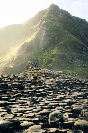 A landscape featuring a formation of interlocking basalt columns against the backdrop of a lush green mountain. The sunlight softly illuminates the mountainous terrain, creating a serene and majestic atmosphere. The focus is on the unique geometric structures of the basalt columns.