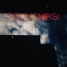 A neon sign reading 'SOUVENIRS!' is visible against a dark reflective glass surface with cloudy skies mirrored on it, creating a moody, industrial atmosphere.