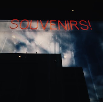 A neon sign reading 'SOUVENIRS!' is visible against a dark reflective glass surface with cloudy skies mirrored on it, creating a moody, industrial atmosphere.